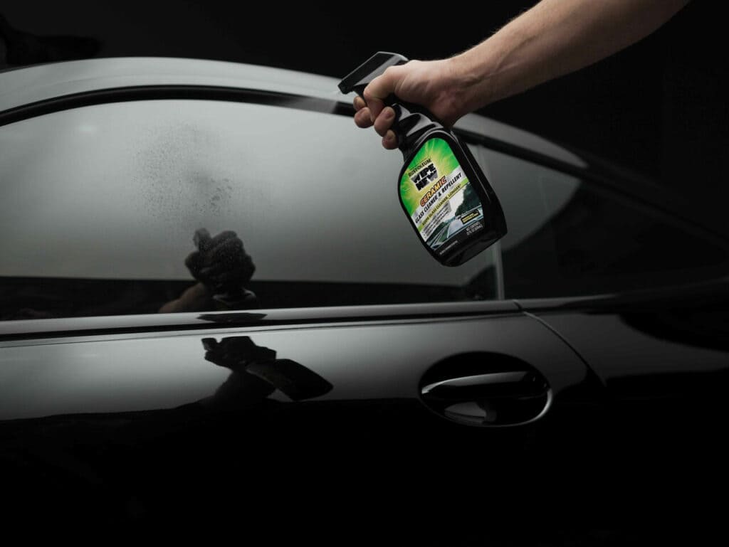 A person spraying a black car with a ceramic auto glass cleaner