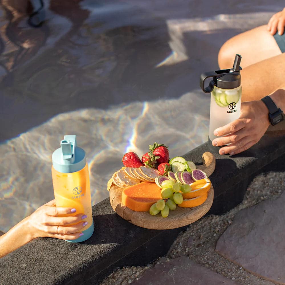 Two hands holdingTakeya glass water bottle with a fruit plate in the middle