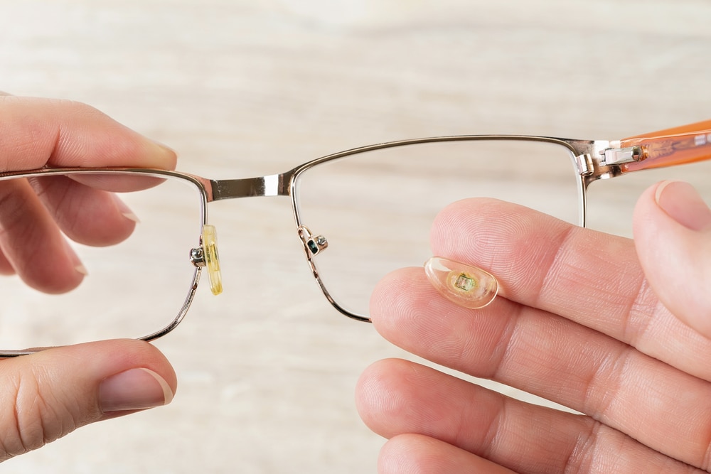 Hands hold eyeglasses and nose pad broke off them against brown desk