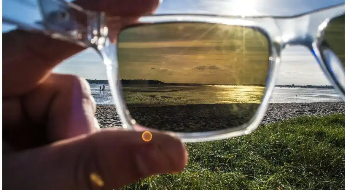 A person holding sunglasses with polarized lenses showing a clear view of the beach and reflecting sunlight in the lenses