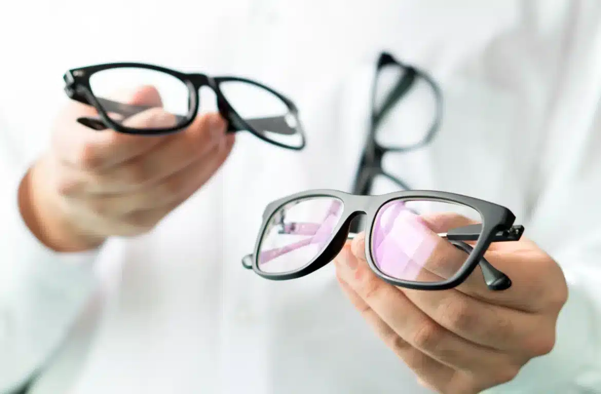 A person holding two pairs of eyeglasses one with clear lenses and the other with a slight purple tint showcasing different styles